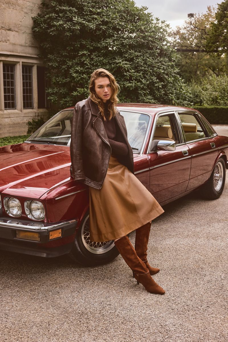 Adult female model poses next to a classic red sedan with a leather jacket, skirt, and suede boots outside a stone building with green foliage.