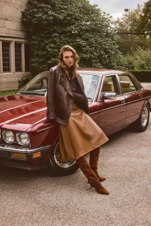 Adult female model poses next to a classic red sedan with a leather jacket, skirt, and suede boots outside a stone building with green foliage.