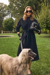 Model in dark coat and sunglasses standing on a lawn holding a dog on a leash for Veronica Beard Resort 2025