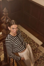 Model wearing a black and white striped cardigan and white trousers leaning on a wooden staircase.