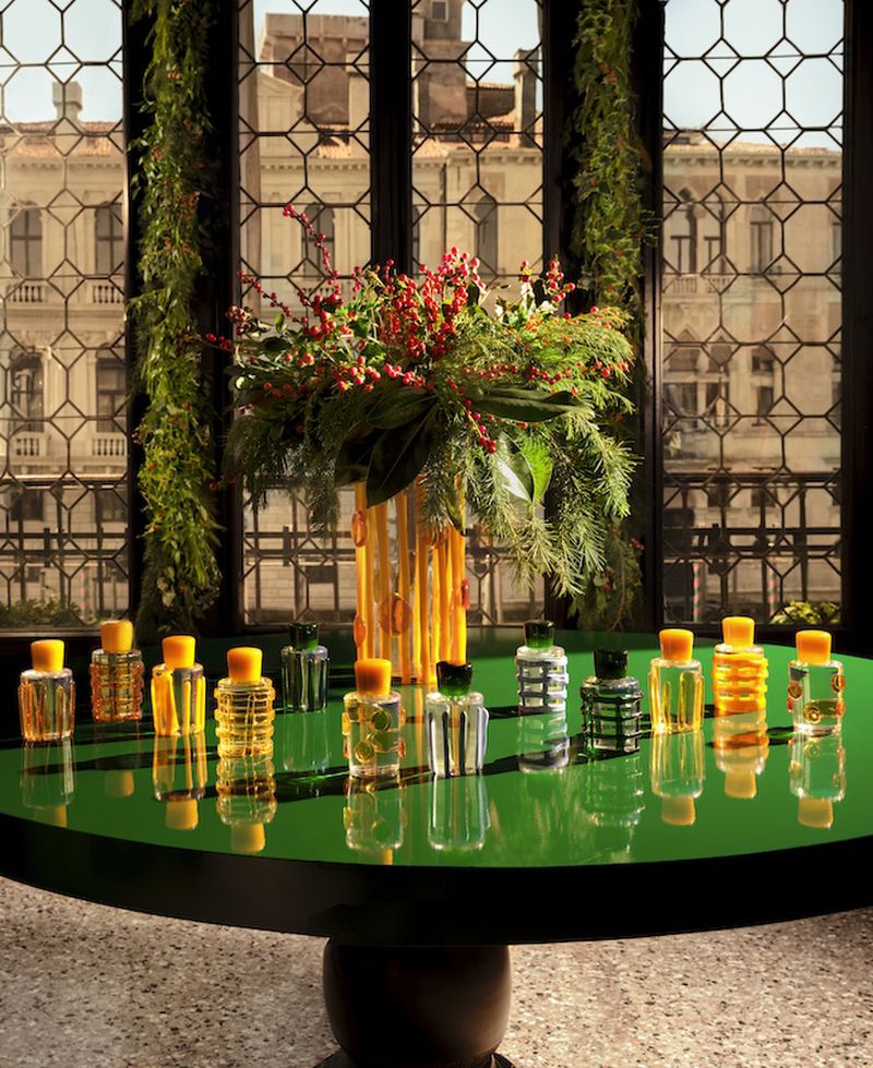 Multiple perfume bottles displayed on a green round table with floral arrangement centerpiece and a window in the background.