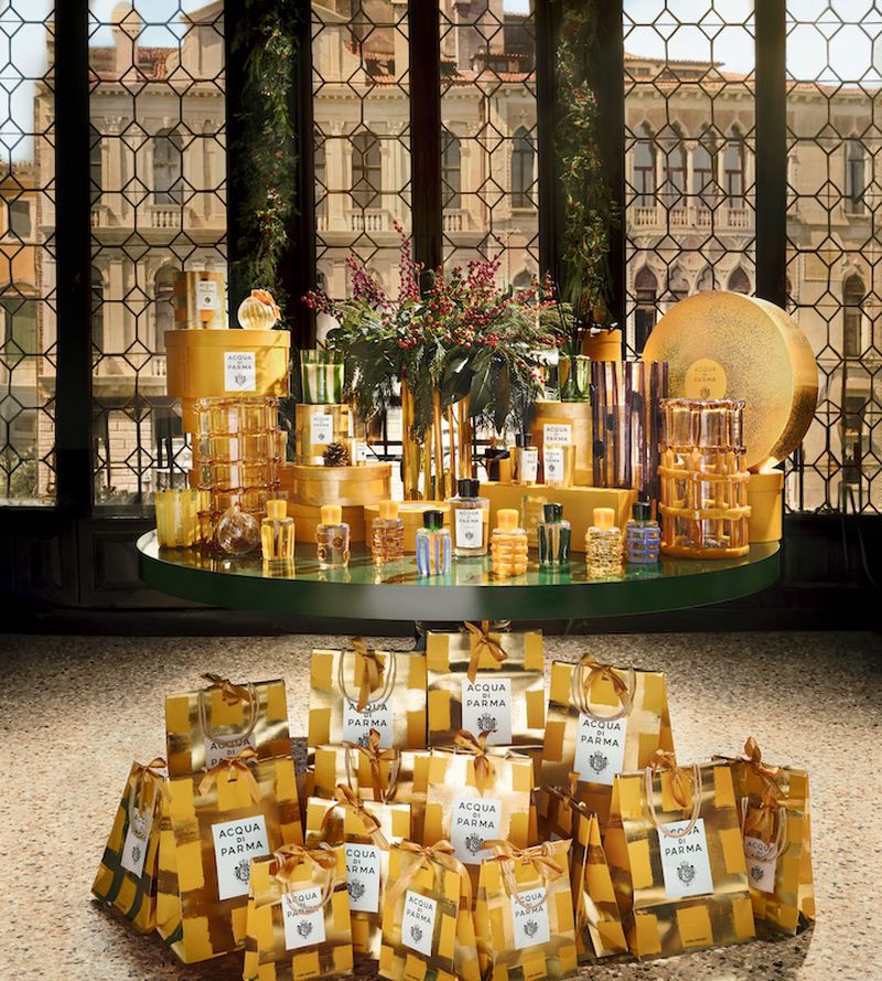 Acqua di Parma product display with gold gift bags and fragrance bottles, set against a backdrop of a building's architectural windows.