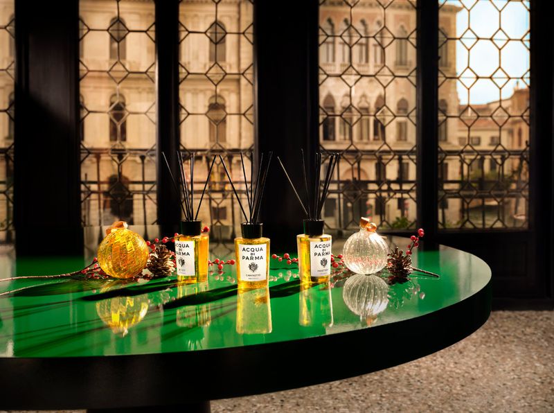 Three Acqua di Parma diffusers are displayed on a glossy green table in front of a window showcasing an Italian building's facade.