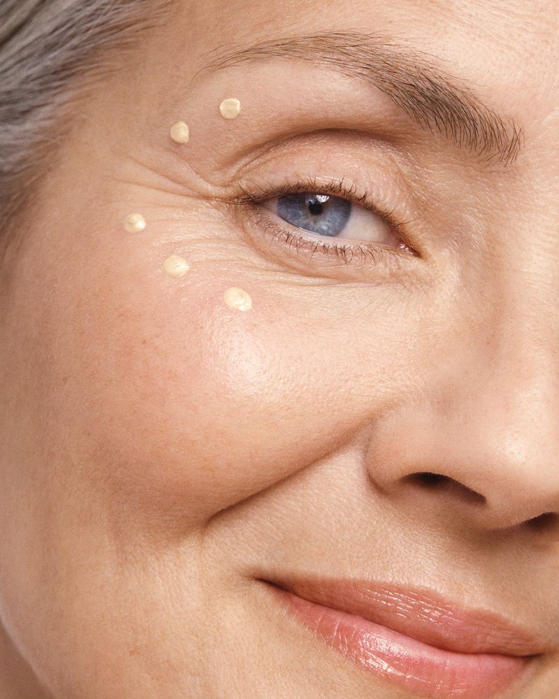 Close-up of an older woman's face, showing her eye, cheek, and lips. She has light skin, grey hair, and blue eyes with a smiling expression.