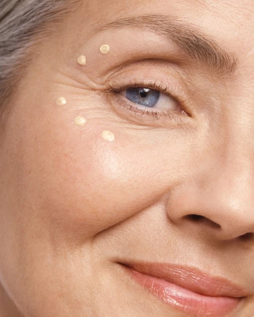Close-up of an older woman's face, showing her eye, cheek, and lips. She has light skin, grey hair, and blue eyes with a smiling expression.