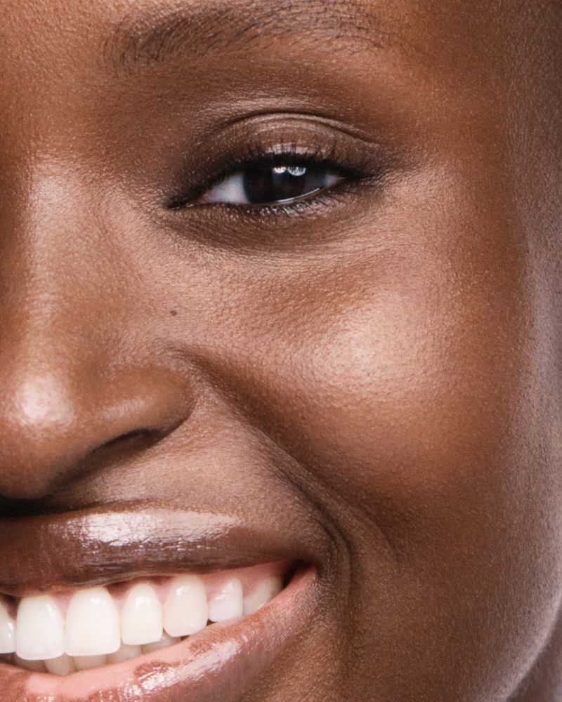 Close-up portrait of a dark-skinned woman featuring her eye, nose, cheek, teeth, and mouth in a beauty or skincare context.