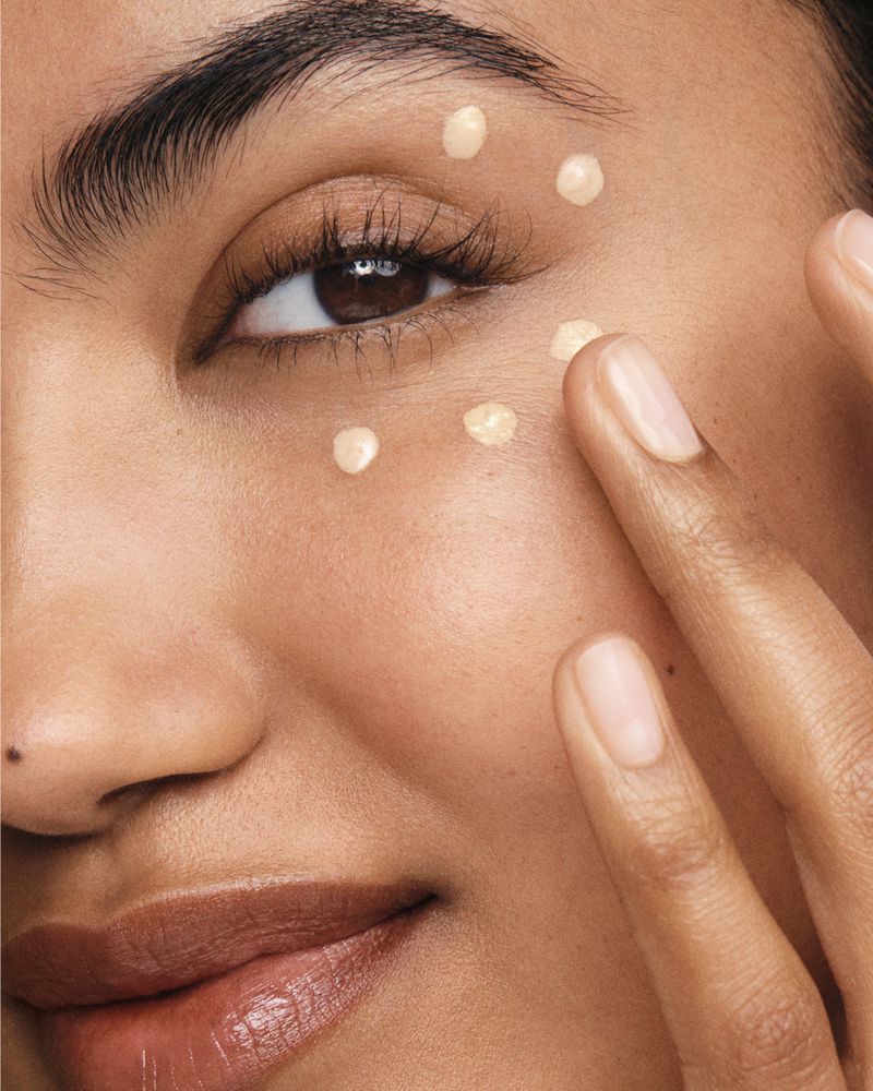 Close-up of woman's face applying small dots of cosmetic product under her eye with her fingertips, showcasing skincare or makeup.