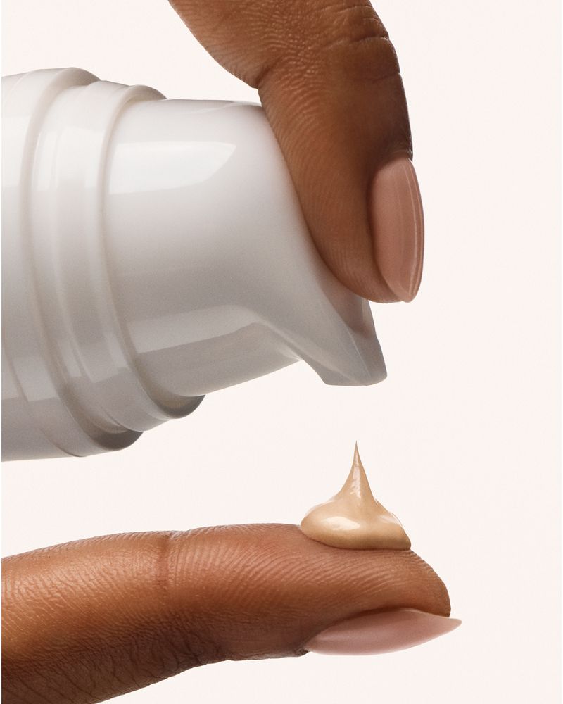 Close-up of a dark-skinned hand with a finger under a white bottle pump, dispensing beige-tinted cream onto the finger nail.
