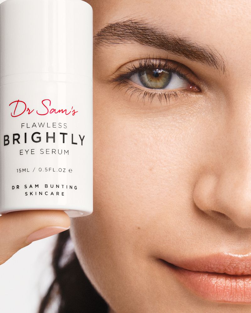 Close-up of a fair-skinned woman's face next to a bottle of Dr. Sam's Flawless Brightly Eye Serum, showcasing skincare product.