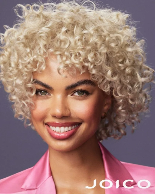 Portrait of a woman with curly blonde hair smiling in front of a dark blue background while wearing a pink jacket in a studio.