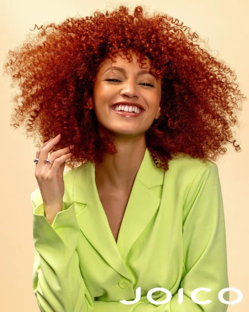 Close-up of a smiling woman with curly red hair, wearing a bright green blazer. Light peach background and Joico logo at the bottom.