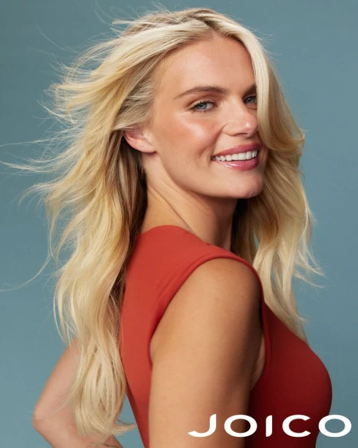 Smiling blonde woman in a sleeveless dress poses against a blue background, showcasing her radiant skin and healthy blonde hair.