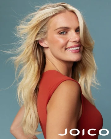 Smiling blond female model in a red sleeveless top with long wind-blown hair against a solid blue background for Joico