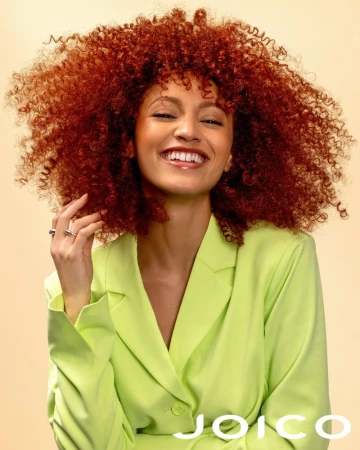 Smiling model with voluminous red curly hair wearing a lime green blazer against a warm neutral background