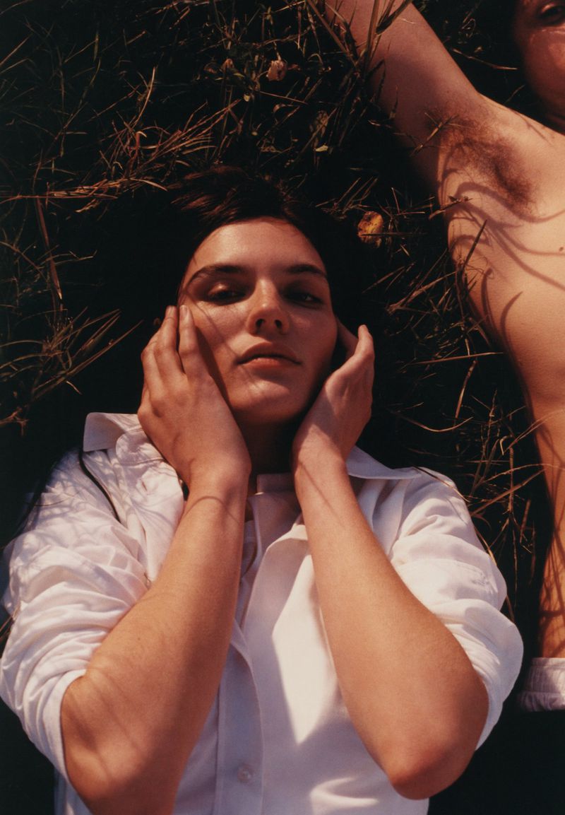Woman lying in grass, hands on face, relaxed mood, next to another person with a sunlit field-like background, casual wear, sunny day.