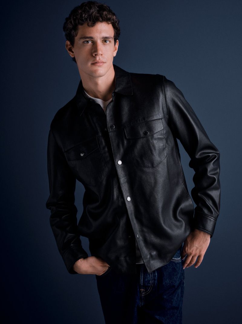 Portrait of a young adult male wearing a black leather shirt and jeans against a dark blue background in a studio setting.