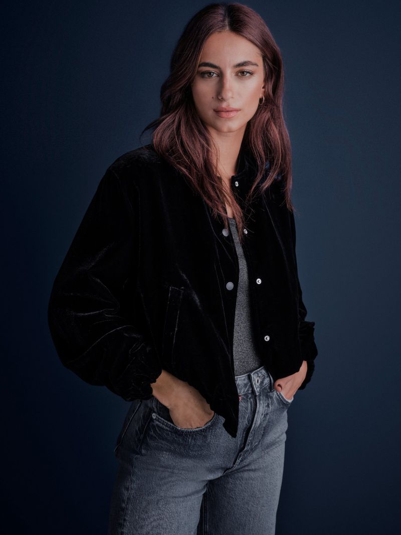 A woman in a black velvet jacket and jeans poses indoors against a dark blue background in a medium shot.