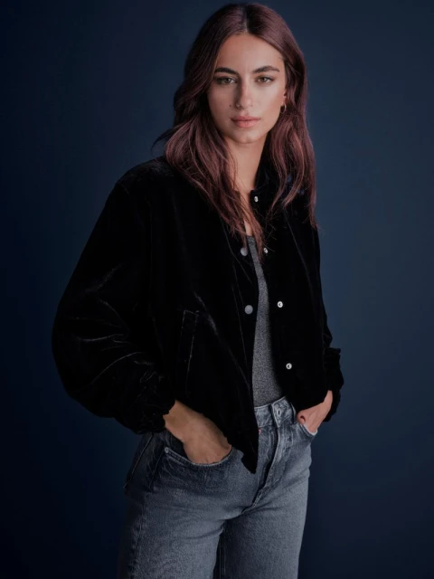 A woman in a black velvet jacket and jeans poses indoors against a dark blue background in a medium shot.