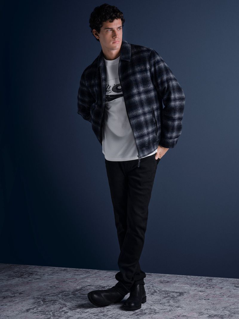 Full-length portrait of a male model wearing a plaid jacket over a white graphic t-shirt against a dark backdrop.