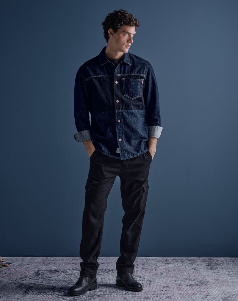 Full-length shot of a young adult male model wearing a blue denim button-down shirt, black pants, and black boots against a blue background.