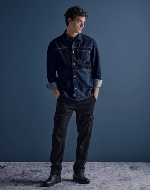 Full-length shot of a young adult male model wearing a blue denim button-down shirt, black pants, and black boots against a blue background.