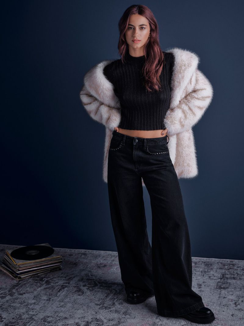 A woman in black jeans, a top, and a faux fur coat stands indoors against a dark blue wall with vinyl records and a textured floor.