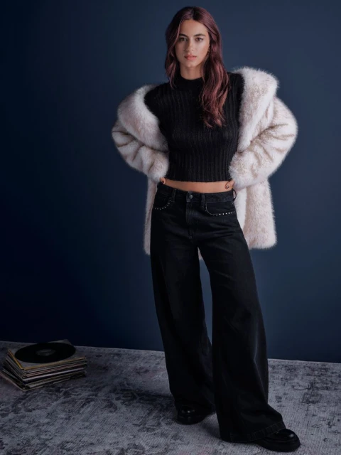 A woman in black jeans, a top, and a faux fur coat stands indoors against a dark blue wall with vinyl records and a textured floor.