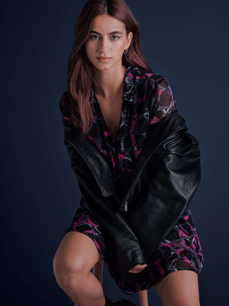 Attractive woman poses in a studio. She wears a printed dress and black jacket, seated against a dark blue background in portrait framing.