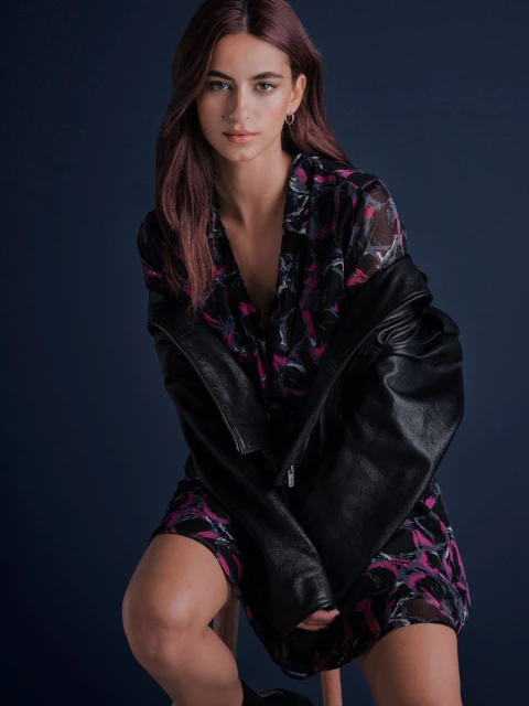 Attractive woman poses in a studio. She wears a printed dress and black jacket, seated against a dark blue background in portrait framing.