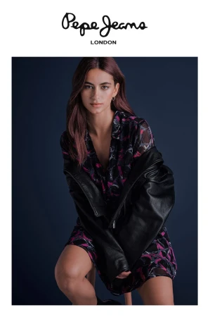 Model in printed dress and leather jacket sitting against dark studio backdrop for Pepe Jeans OI24 campaign.