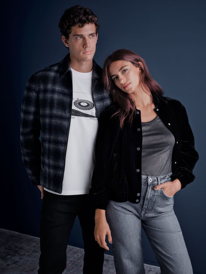 A stylish couple poses together indoors. The man wears a plaid shirt and graphic tee, while the woman sports a velvet jacket and jeans.