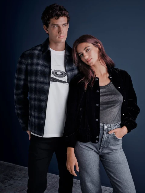 A stylish couple poses together indoors. The man wears a plaid shirt and graphic tee, while the woman sports a velvet jacket and jeans.