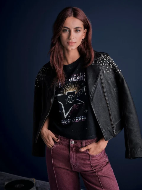 Stylish woman with auburn hair poses in a black studded leather jacket and red jeans against a dark blue backdrop in a studio setting.
