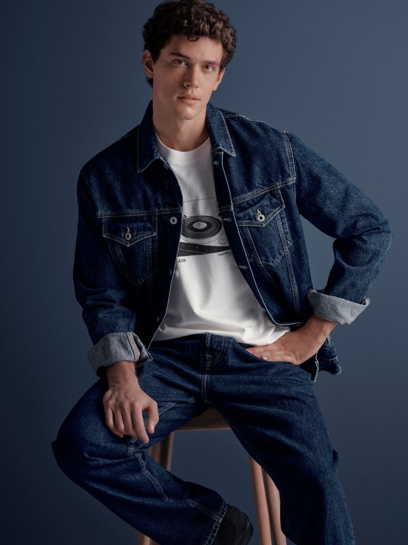 Man in denim jacket and jeans seated on stool. Studio shot with a relaxed pose. Male fashion, denim on denim, studio lighting.