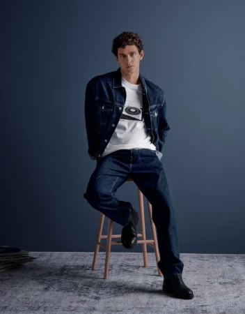 Model Xavier Serrano wearing a denim jacket and jeans seated on a wooden stool against a dark blue background for Pepe Jeans
