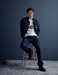 Model Xavier Serrano wearing a denim jacket and jeans seated on a wooden stool against a dark blue background for Pepe Jeans
