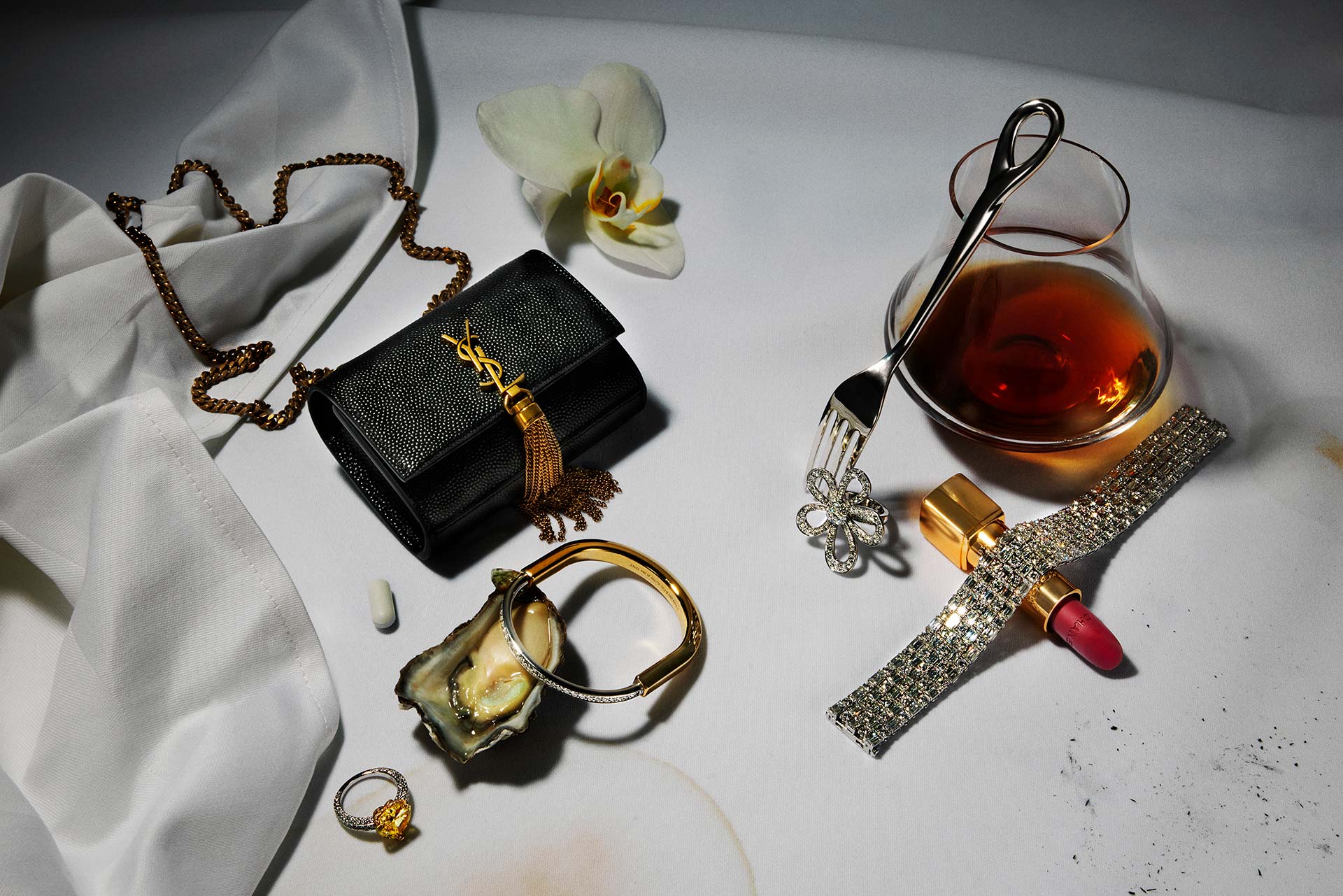 Luxury still life featuring a black YSL purse, jewelry, makeup, oyster shell, white orchid, and glass of amber liquid on white fabric.