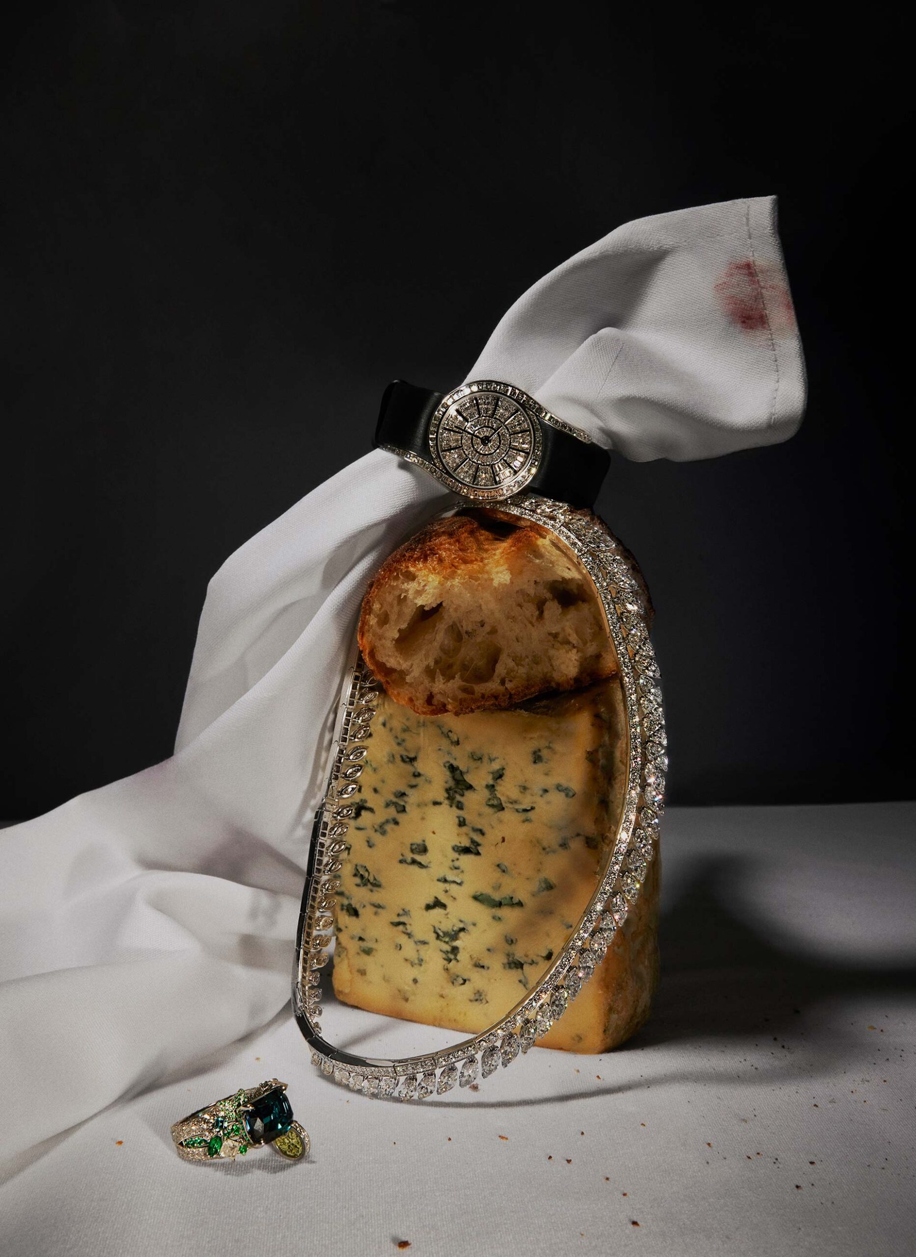 Still life shot of a diamond bracelet, ring and wrist watch on a slice of blue cheese and a white cloth against a dark background.