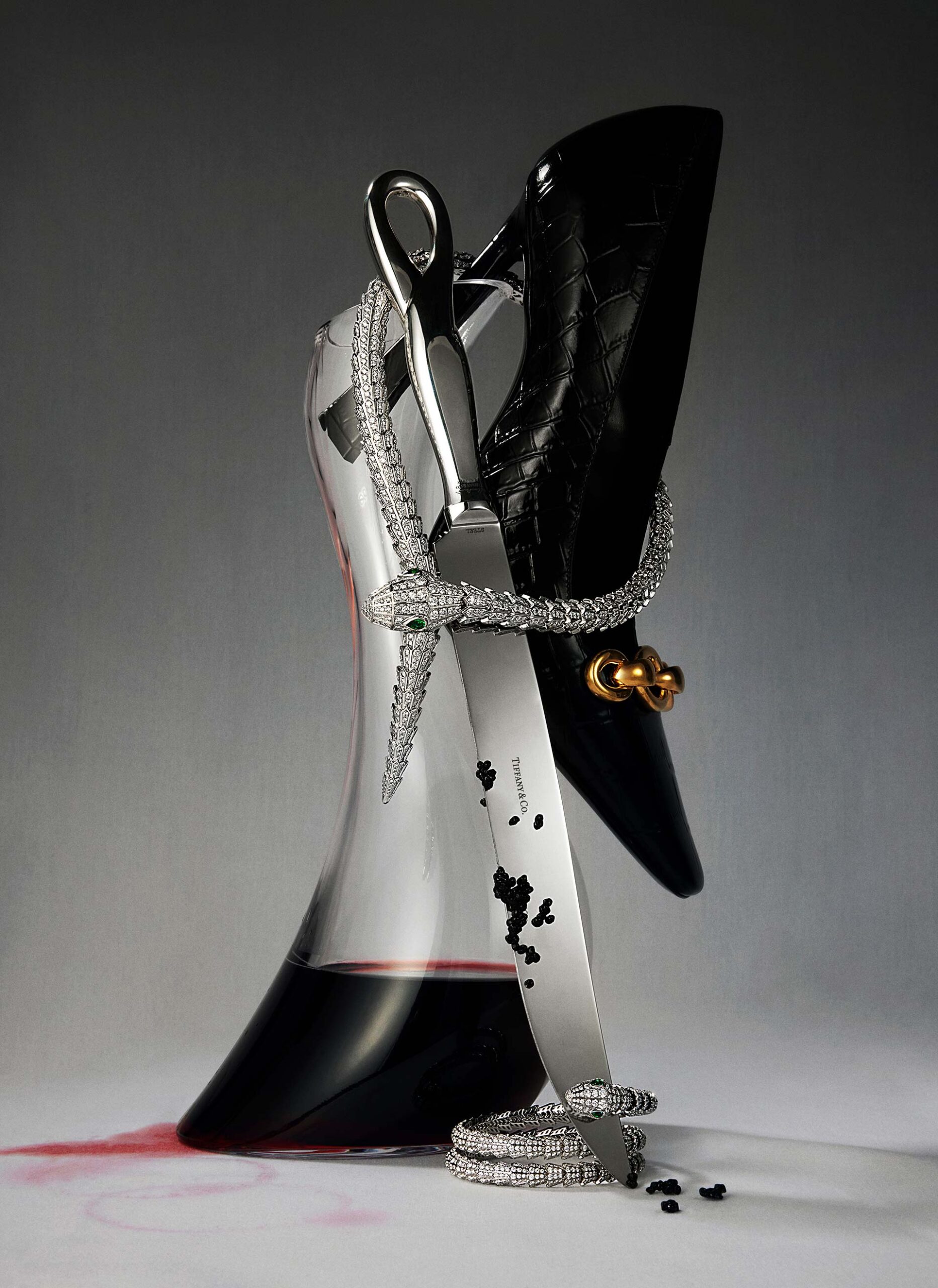Still life shot of a black high heel shoe resting on a crystal wine decanter, a diamond snake necklace, silver flatware, and wine spill.