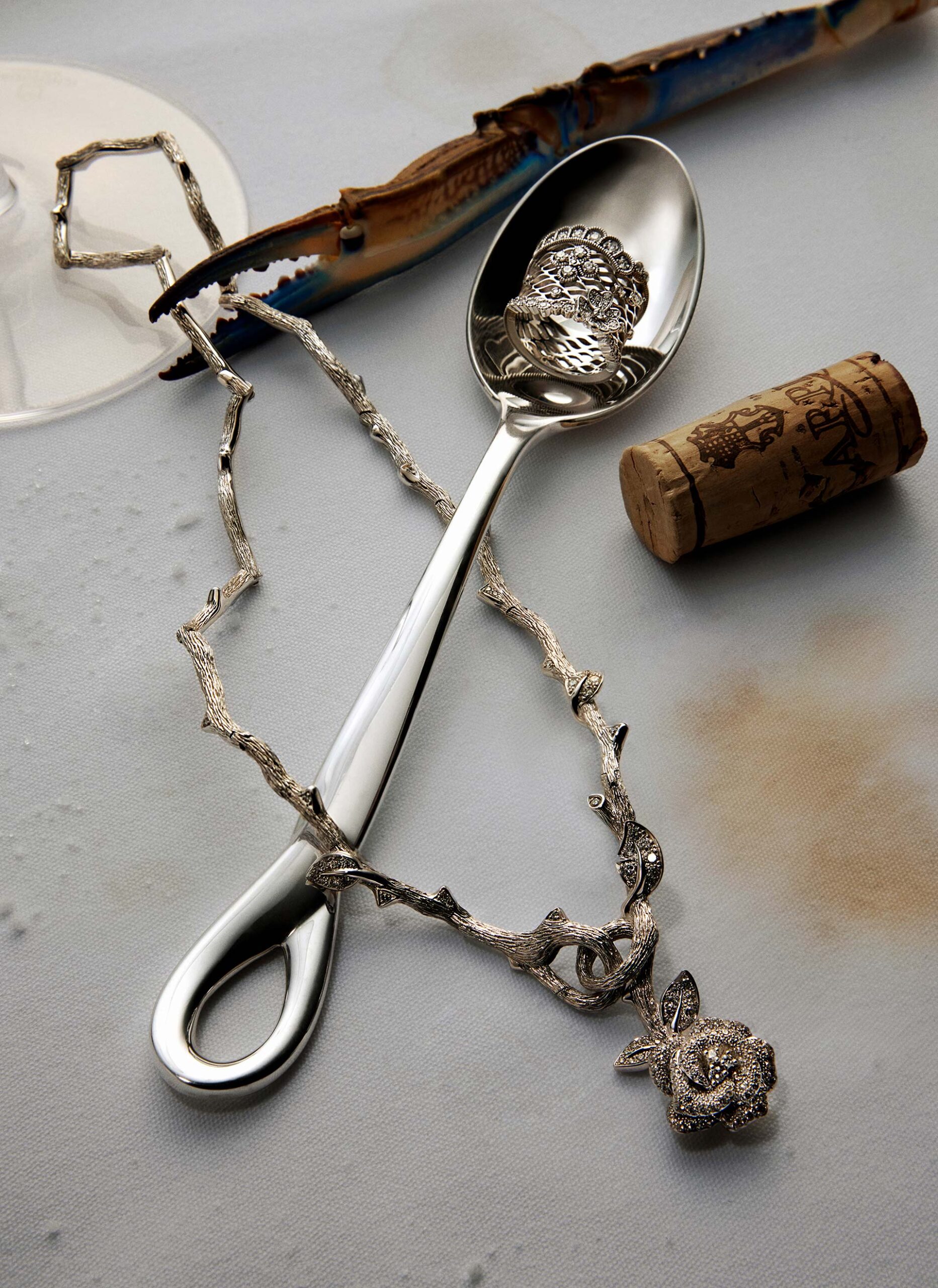 Still life featuring a silver spoon cradling a silver ornamental ring, accompanied by a rose necklace, crab claw, and wine cork on white surface.