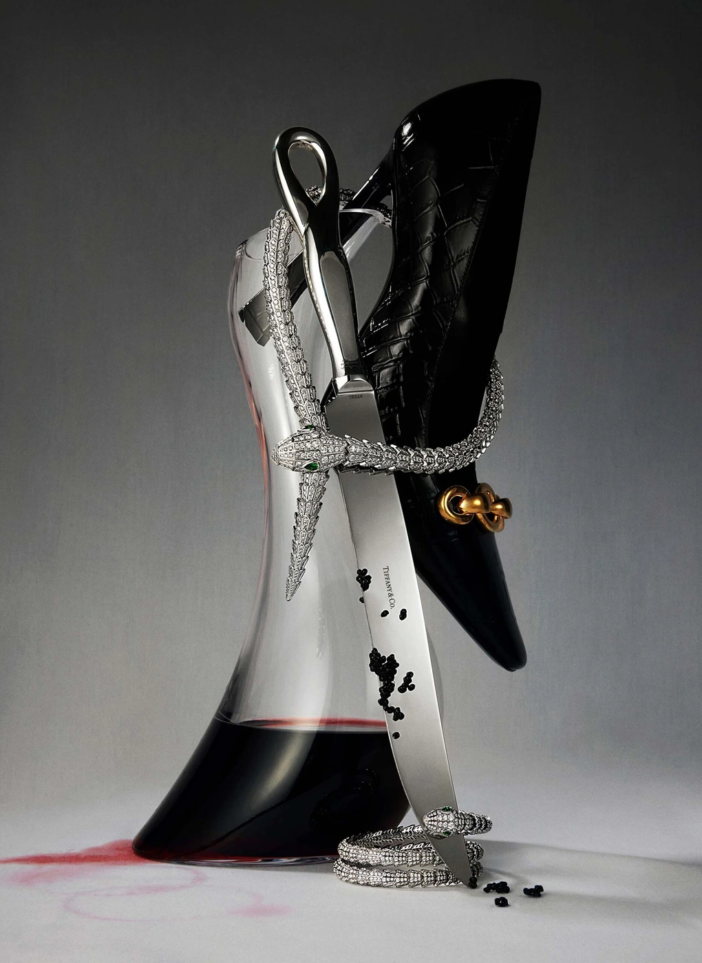 High heel shoe, crystal decanter with wine, silver knife, and diamond snake necklace on a grey surface.
