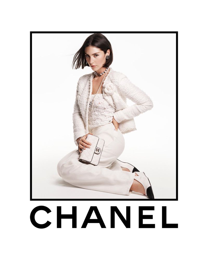 A fashionable adult woman in a light-colored tweed suit, accessorized with jewelry and a handbag, kneels in a studio setting for a portrait.