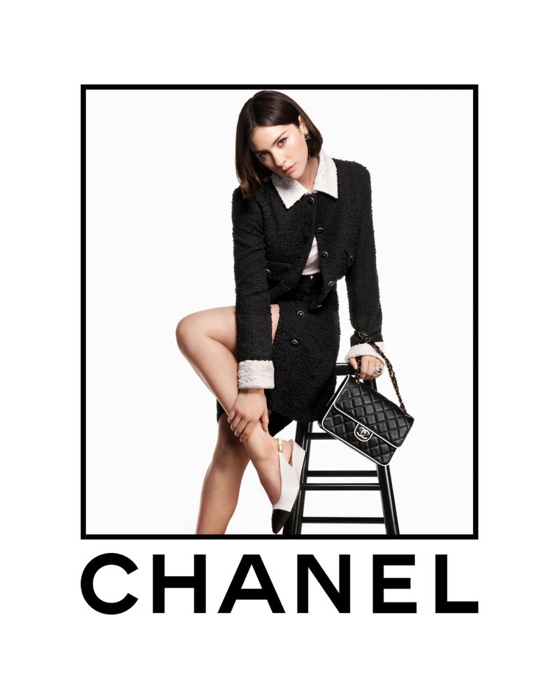 An adult woman poses in a Chanel-branded advertisement, wearing a black jacket and skirt, holding a black purse, and sitting on a stool.