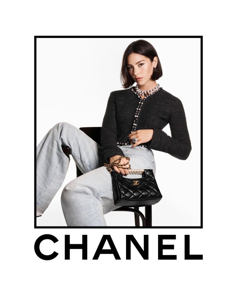 Gracie Abrams wearing a black tweed jacket and light wash jeans holding a black quilted handbag against white