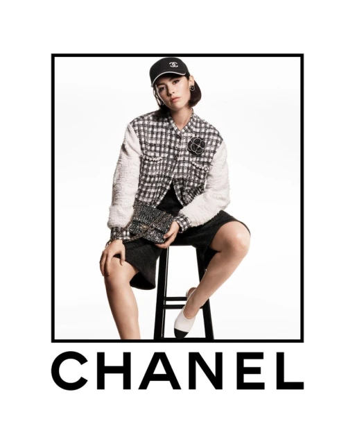 A woman sits on a stool in a fashion studio wearing a Chanel-branded baseball cap, jacket, skirt, and shoes, holding a small handbag.