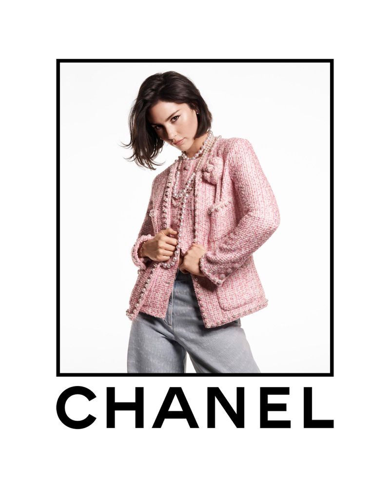 Elegant woman posing in a pink tweed Chanel jacket with light-wash jeans against a white background, highlighted by a black frame.