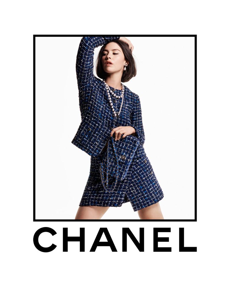 Gracie Abrams wearing a blue tweed Chanel suit and pearl necklace holding a matching handbag against a white background