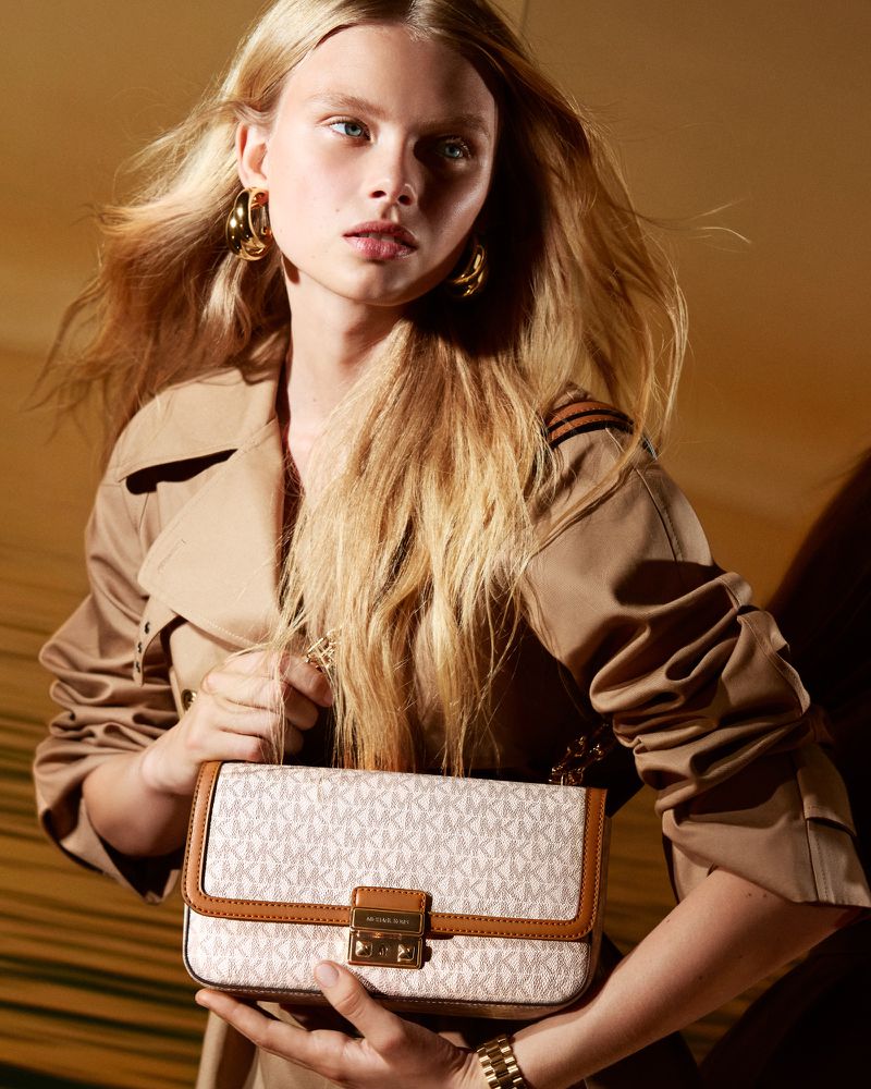 Fashion shot of a blonde woman in a trench coat holding a patterned Michael Kors purse in front of a neutral backdrop.