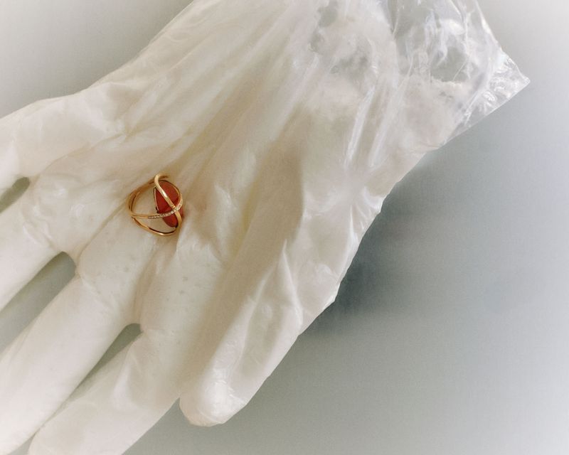 A gold ring with an orange gemstone held in a gloved hand, providing a close-up view of the jewelry against a clean, light background.