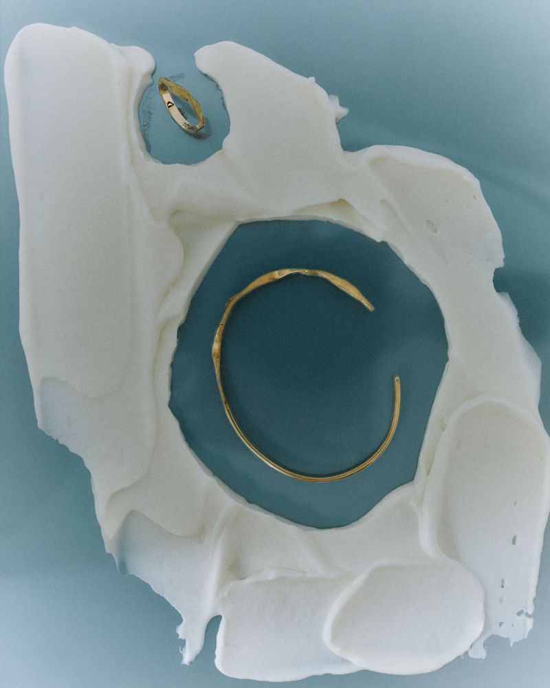 Elevated shot of a gold ring and open bracelet arranged on a textured white surface against a pale blue backdrop, jewelry showcase.
